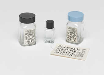 A clear glass bottle with a blue screw lid.  A Maciunas-designed label is attached to the...