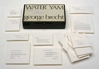 Translucent plastic box with a Maciunas-designed label attached to the lid, containing 98 score...