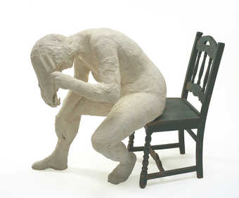 A plaster woman seated on a green wooden chair.  The woman is bent at the waist and holds a...