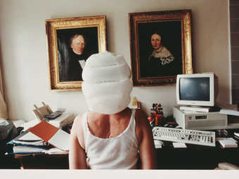 image of the back of a woman from waiste up, with a white bag over her head, sitting at a desk in...