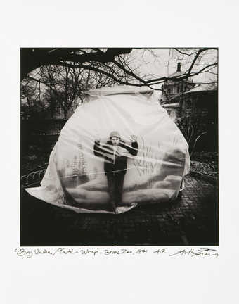 An image of a boy covered with a sheet of plastic, standing in front of a giant fish bowl.