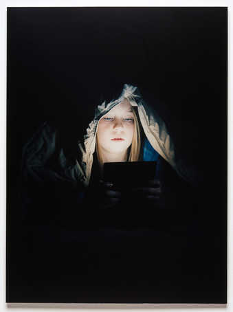 An image of girl with a comforter on her head whose face is illuminated by the electronic device...